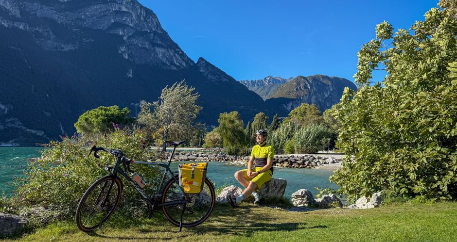 Cyclist in yellow sitting on grass by turquoise lake with touring bike. Mountains and trees frame the scenic lakeside promenade in Riva.
