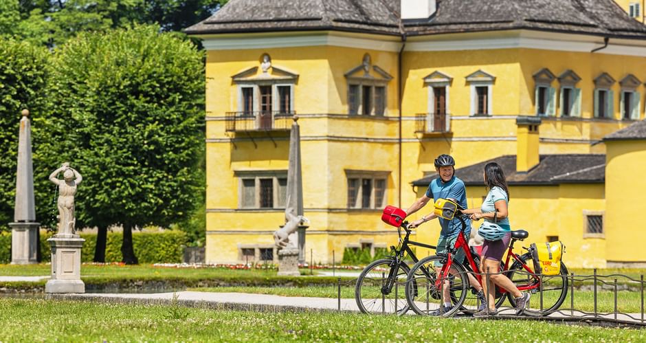 Two cyclists with bicycles on a lawn in front of the yellow Hellbrunn Palace in Salzburg, with statues and green trees visible in the park.