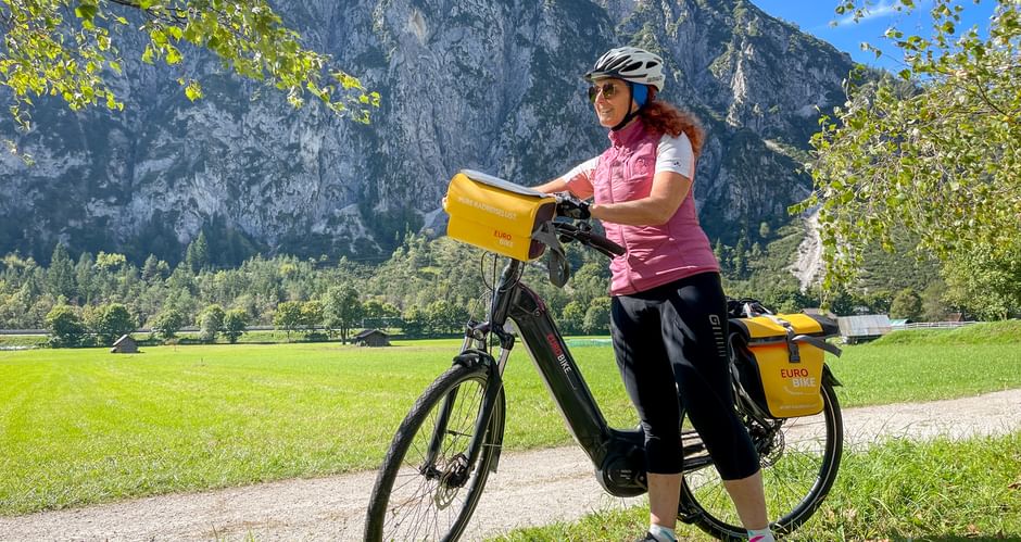 Frau in rosa Radtrikot mit Helm neben E-Bike mit gelben Packtaschen. Felsige Berge und grüne Wiese im Hintergrund.