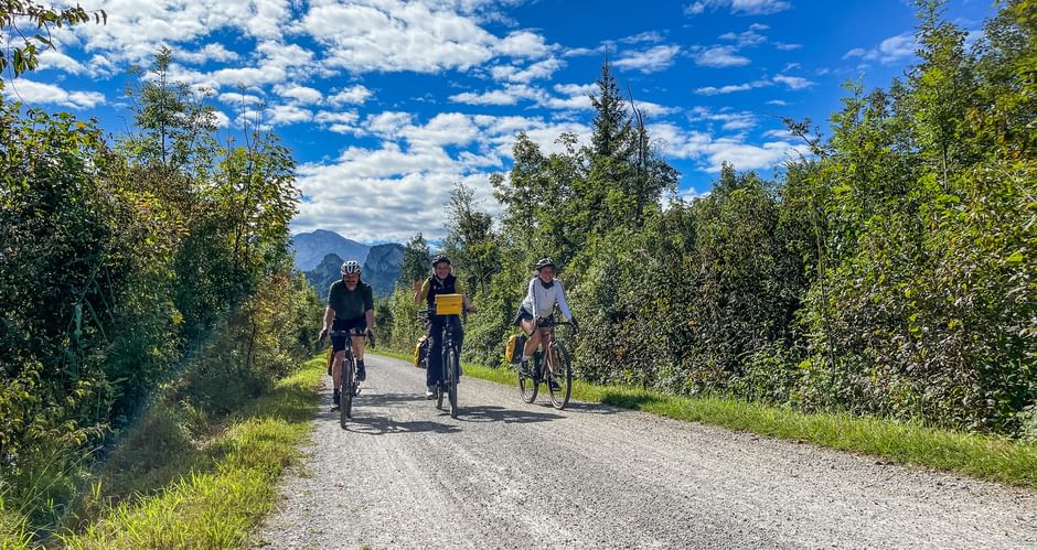 Three cyclists riding on a gravel path along the Salzach cycle route near Rif, surrounded by green trees with mountains in the background.