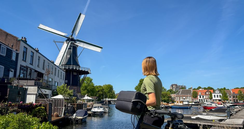 Radfahrerin mit Fahrrad betrachtet die historische Windmühle De Adriaan am Fluss Spaarne in Haarlem, mit Booten und traditionellen Gebäuden.