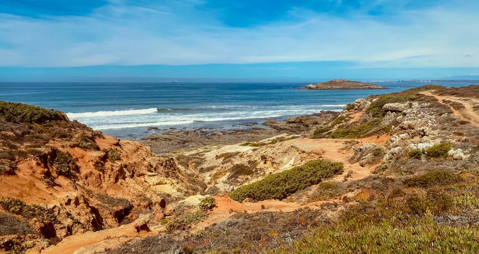 Zerklüftete Küstenlandschaft bei Porto Covo mit rotbraunen Klippen, grüner Vegetation und blauem Atlantik mit weißen Wellen und kleinen Inseln.