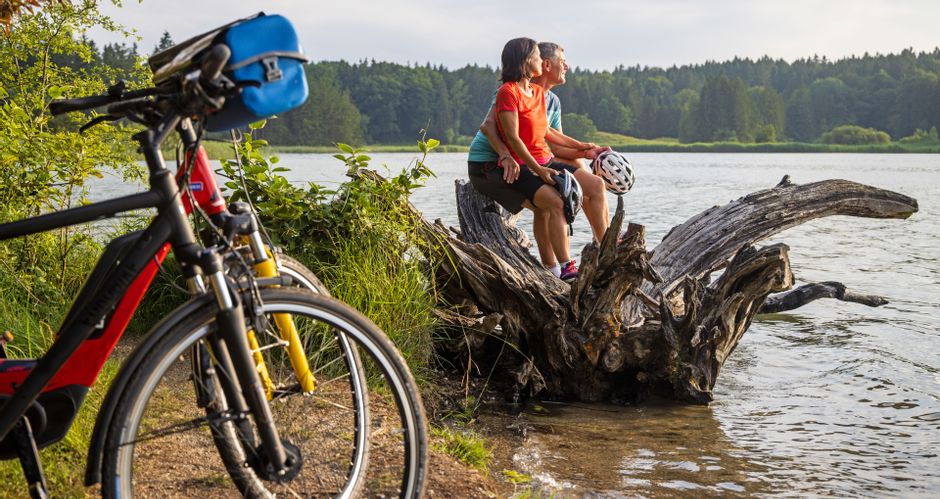 Cycling break of a couple sitting on a large root on the shore of Lake Ostersee
