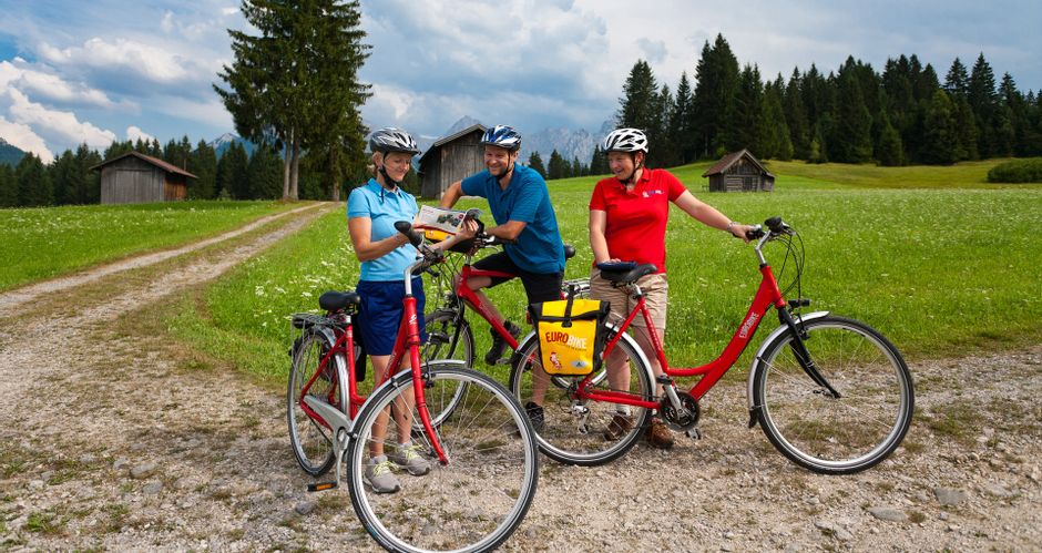 Eine Gruppe von Radfahrern bei einem Blick in die Karte, im Hintergrund Wiesen, Heuschober, Wald und Berge