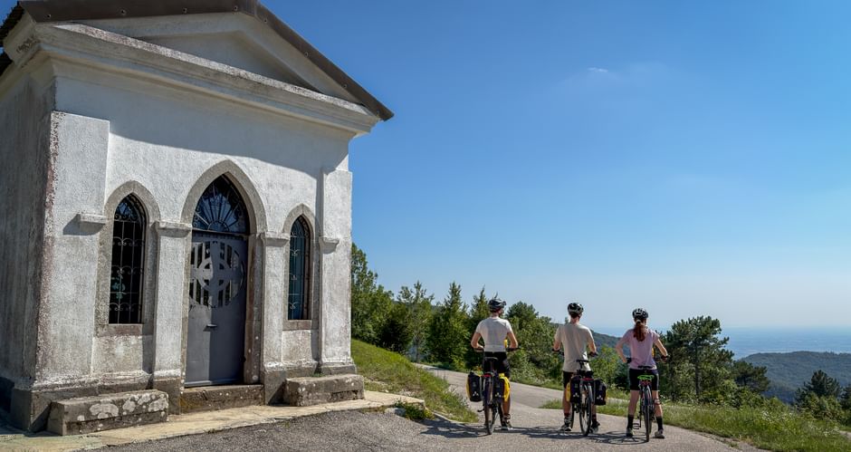 Three cyclists on a hilltop road beside a small white chapel with Gothic windows near Canebola, overlooking forested hills and distant sea.