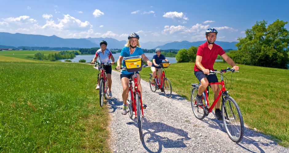 A group of cyclists on a gravel cycle path across meadows, with Lake Rieg and the mountains in the background
