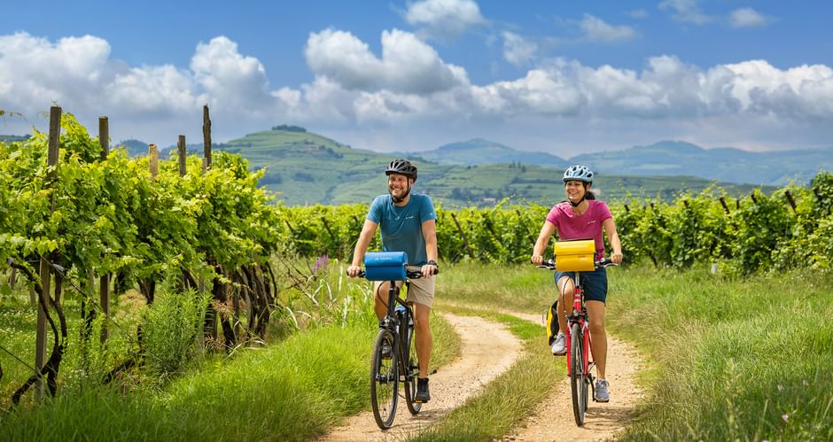 Zwei Radfahrer auf einem Feldweg zwischen grünen Weinreben unter blauem Himmel mit sanften Hügeln in der Prosecco-Region.