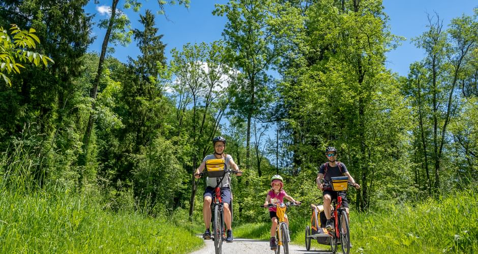 Family of four cycling on gravel path along Lechradweg. Two adults and two children with bike trailer ride through green landscape under blue sky.