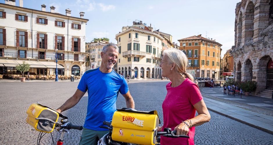 Zwei Radfahrer in der Innenstadt von Verona, im Hintergrund die Arena und typisch italienische Häuser