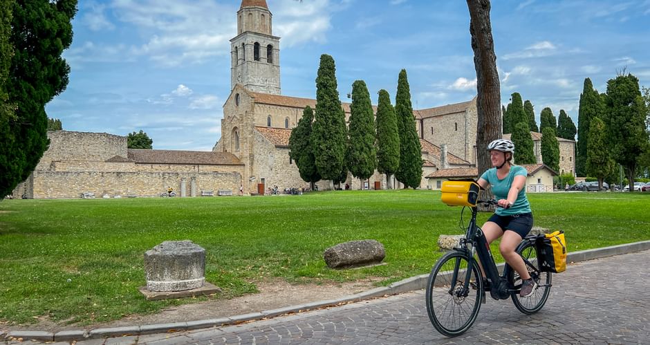 Radfahrerin mit gelben Taschen fährt an der Basilika Santa Maria Assunta in Aquileia vorbei. Steinkirche mit Glockenturm und Zypressen sichtbar.