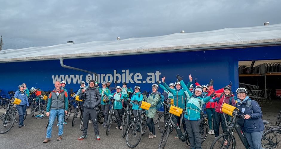 Large group of cyclists with bikes and yellow bags posing in front of blue Eurobike building during company trip along the Danube.