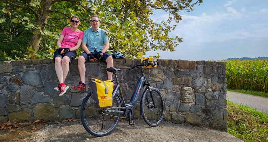 Zwei Radfahrer mit Sonnenbrillen sitzen auf einer Steinmauer mit ihrem geparkten Fahrrad davor, am Alpe Adria Radweg bei Grado.