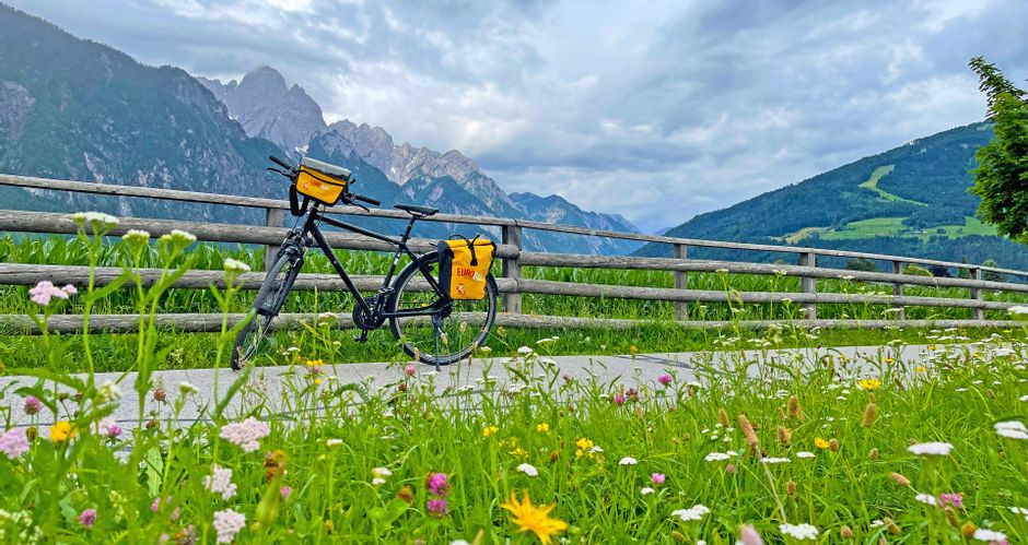 Ein Fahrrad steht vor einem Weidezaun mit Blumenwiese im Vordergrund und den Lienzer Dolomiten im Hintergrund