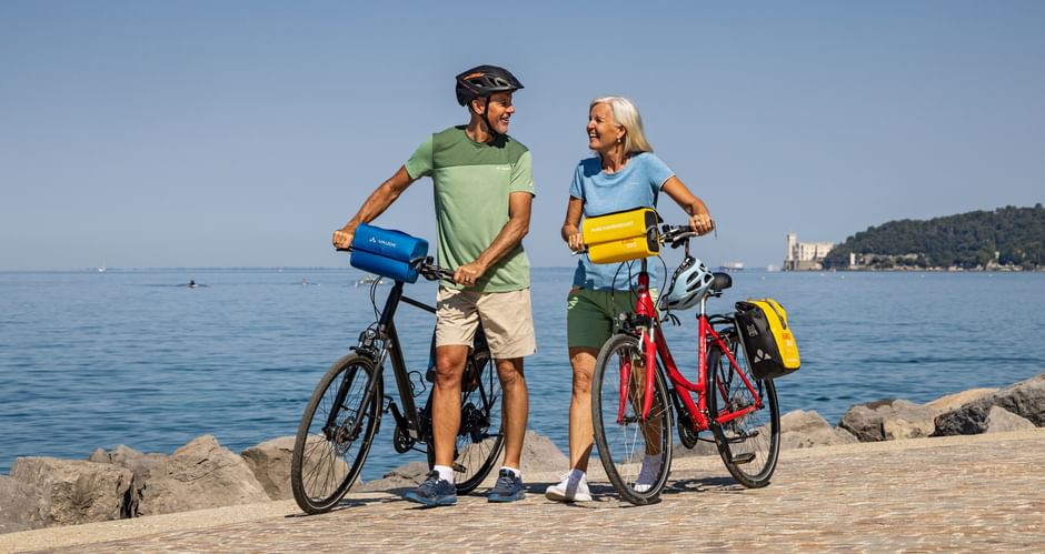 Paar mit Tourenrädern auf gepflastertem Uferweg bei Triest. Schloss Miramare auf entferntem Hügel über ruhiger blauer Adria sichtbar.