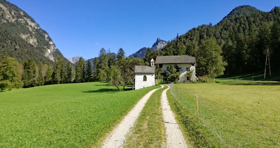 Kleine weiße Kapelle und alter Bauernhof am Wegesrand, mit Bergen und Wald im Hintergrund
