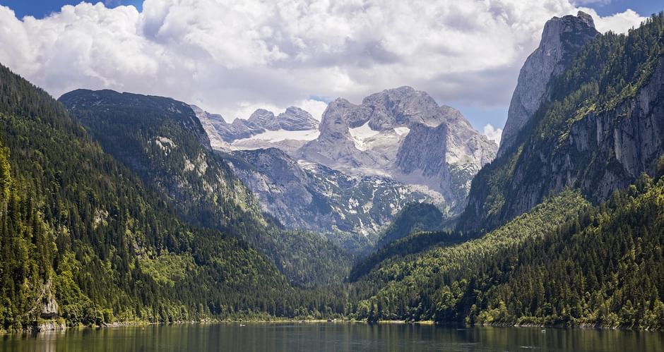 Lake Gosausee in Salzkammergut with forested mountains and Dachstein massif with snow-capped peaks under dramatic clouds.