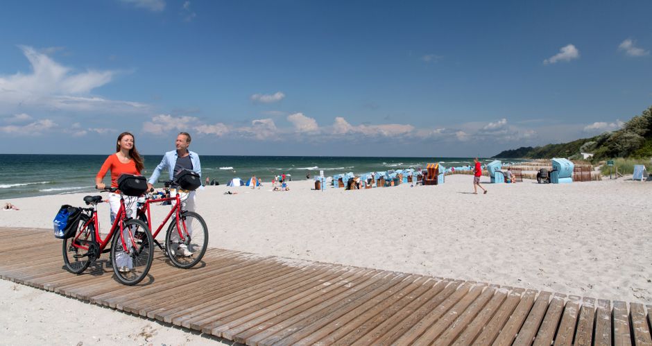Zwei Radfahrer schieben ihrer Räder auf einem Holzsteg am Strand, im Hintergrund Strandkörbe
