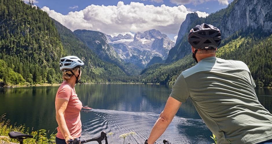 Two cyclists with helmets pause at Gosausee lake shore, viewing the Dachstein mountain range with snow-capped peaks and forested slopes.