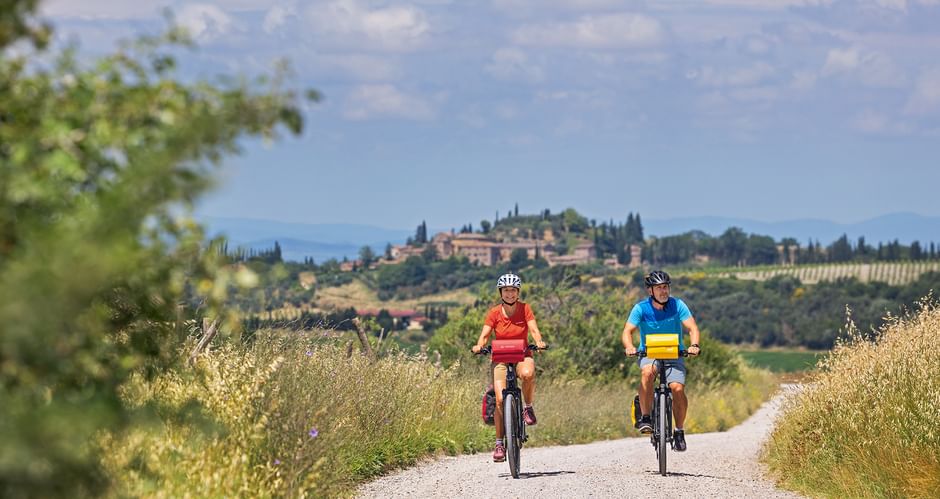 Zwei Radfahrer auf einem Schotterweg durch goldene Felder in der Toskana Val d'Orcia, mit einem Bergdorf und Zypressen im Hintergrund.