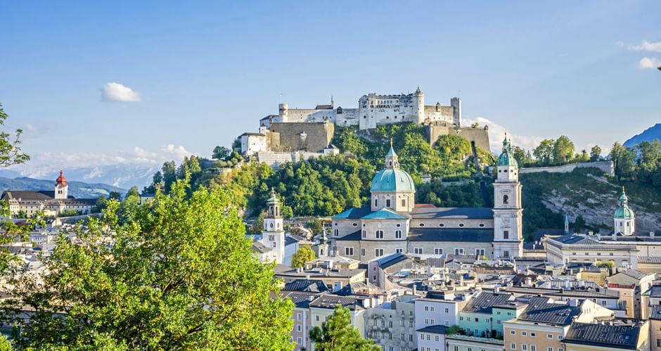 Salzburg cityscape with historic buildings, baroque church domes, and Hohensalzburg Fortress on a hilltop. Green trees frame the view under blue sky.