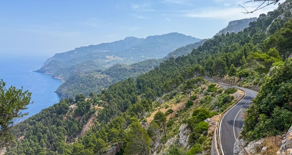 Kurvenreiche Küstenstraße durch das Tramuntana-Gebirge auf Mallorca mit blauem Mittelmeer und bewaldeten Hängen unter klarem Himmel.
