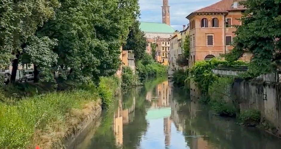 Canal in Vicenza with red flowers in foreground, historic buildings and green copper-roofed church with tall bell tower reflected in water.