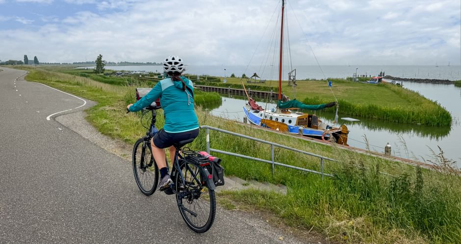 Female cyclist in turquoise jacket riding on paved dike path along IJsselmeer with traditional sailboat moored in canal and green marshland.