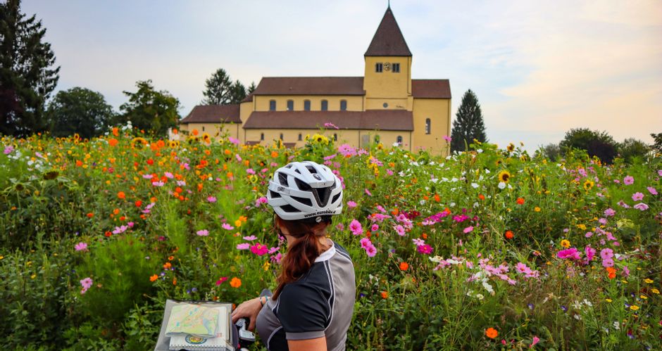 A cyclist stands in front of a flower meadow and a church