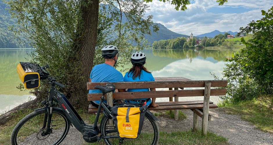 Radlerpaar sitzt auf Bankerl und blickt auf Weissensee
