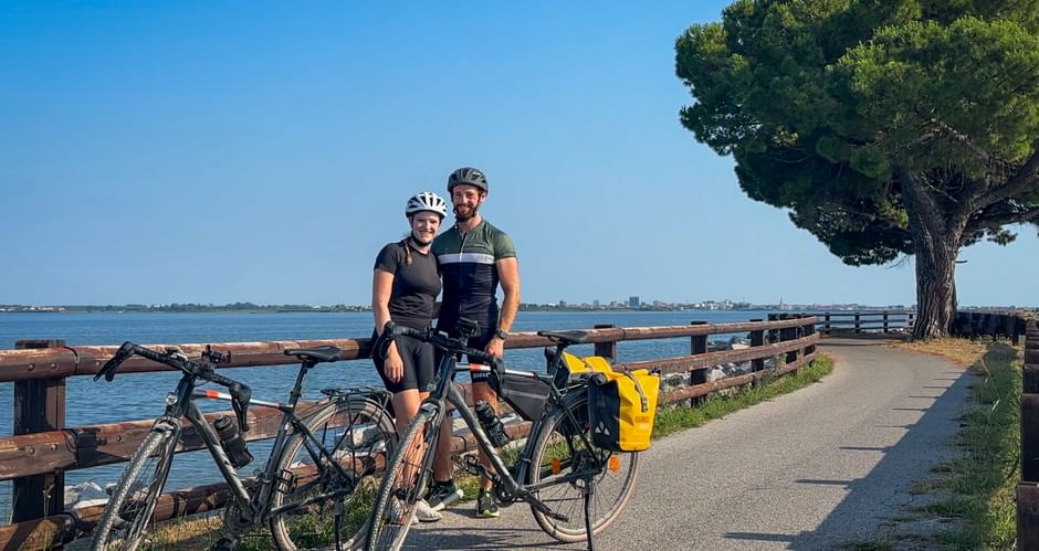 Two cyclists with helmets and touring bikes on coastal path with wooden fence, lagoon, and pine tree near Grado on the Alpe Adria cycle route.