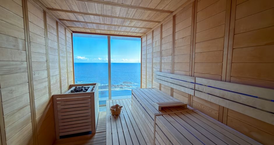 Light wood sauna interior with tiered benches and heater, featuring large window overlooking blue ocean in Porto Moniz, Madeira.