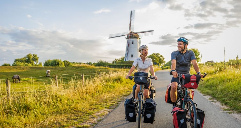 Zwei Radfahrer mit Packtaschen fahren auf einer asphaltierten Landstraße in Flandern. Eine weiße Windmühle steht im Hintergrund unter bewölktem Himmel.
