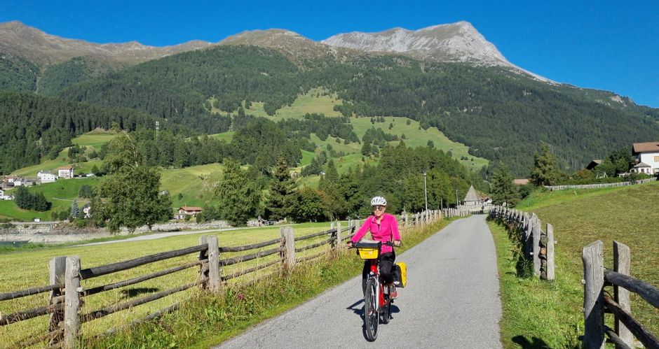 Cyclist on asphalted cycle path surrounded by fenced meadows, mountains in the background