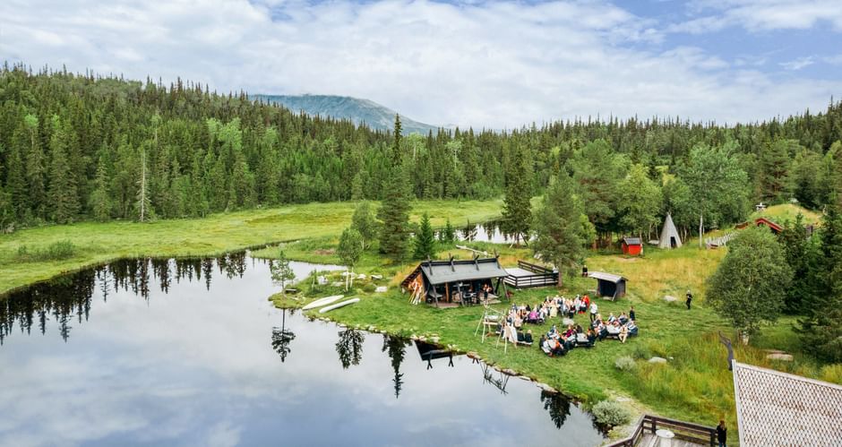 Luftaufnahme einer Versammlung am ruhigen See in Hemsedal Kommune. Menschen sitzen um eine Holzhütte, umgeben von grünem Wald und Bergen.