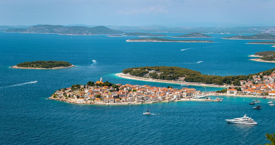 Aerial view of Primošten peninsula with terracotta roofs, church tower, and turquoise Adriatic Sea. Islands and mountains visible in background.