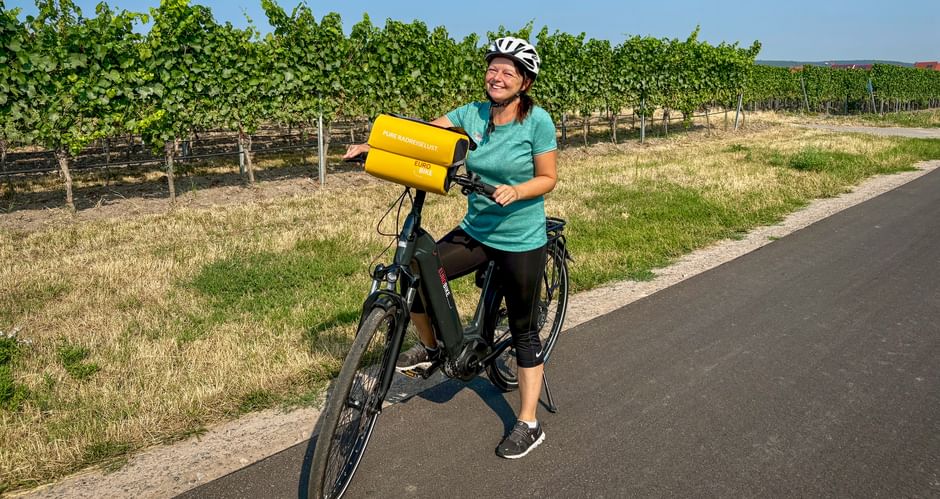 Radfahrerin mit Helm hält gelbe Fahrradtasche auf Radweg neben Weinbergreihen unter blauem Himmel in der Region Speyer-Heidelberg-Worms.