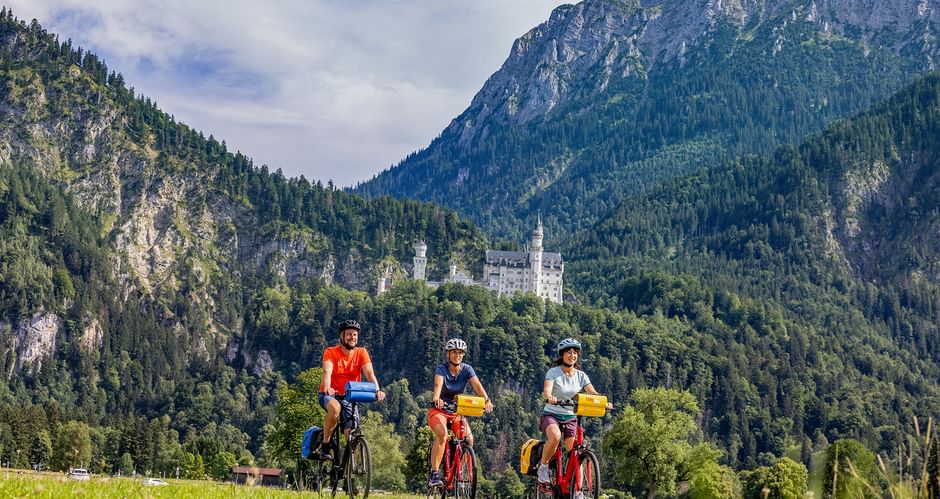 Drei Radfahrer fahren durch eine grüne Wiese mit Schloss Neuschwanstein und bewaldeten Bergen im Hintergrund unter blauem Himmel.