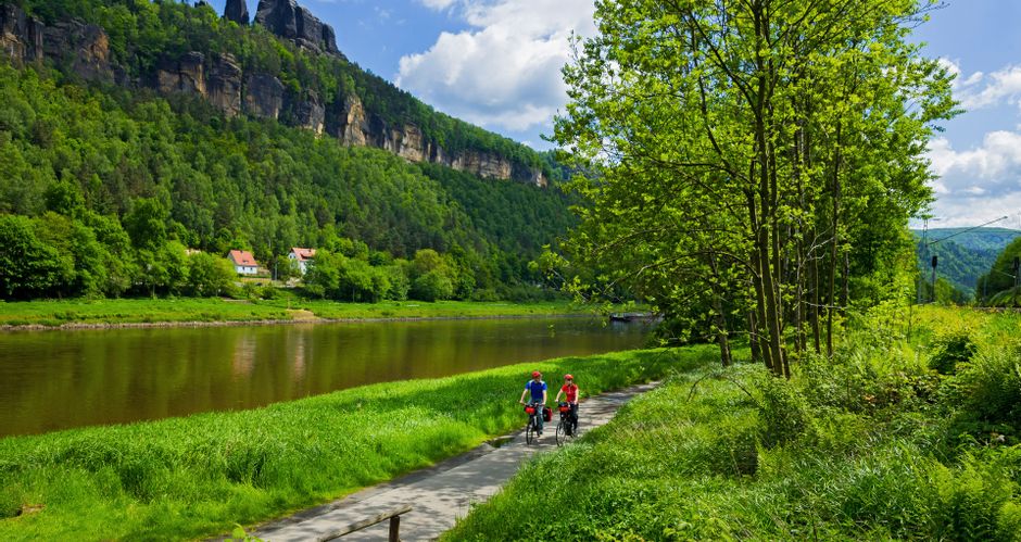 Zwei Radfahrer fahren entlang der Elbe, im Hintergrund Felsen und Wälder