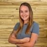 Vanessa Kainz mit langen braun-blonden Haaren und grauem Poloshirt, steht mit verschränkten Armen lächelnd vor einer Holzwand.