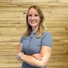 Stefanie Kastenauer smiling in a blue polo shirt, standing with crossed arms in front of a light wooden wall.