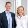 Thomas Schmid in dark blazer and light blue shirt with Verena Sonnenberg in white blouse, both smiling in a bright office setting.